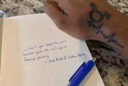 The image shows an open noted book with writing on it, a blue pen laying across the page and a hand above both which is tattooed with female and male gender signs, and a heart monitor reading leading out from them