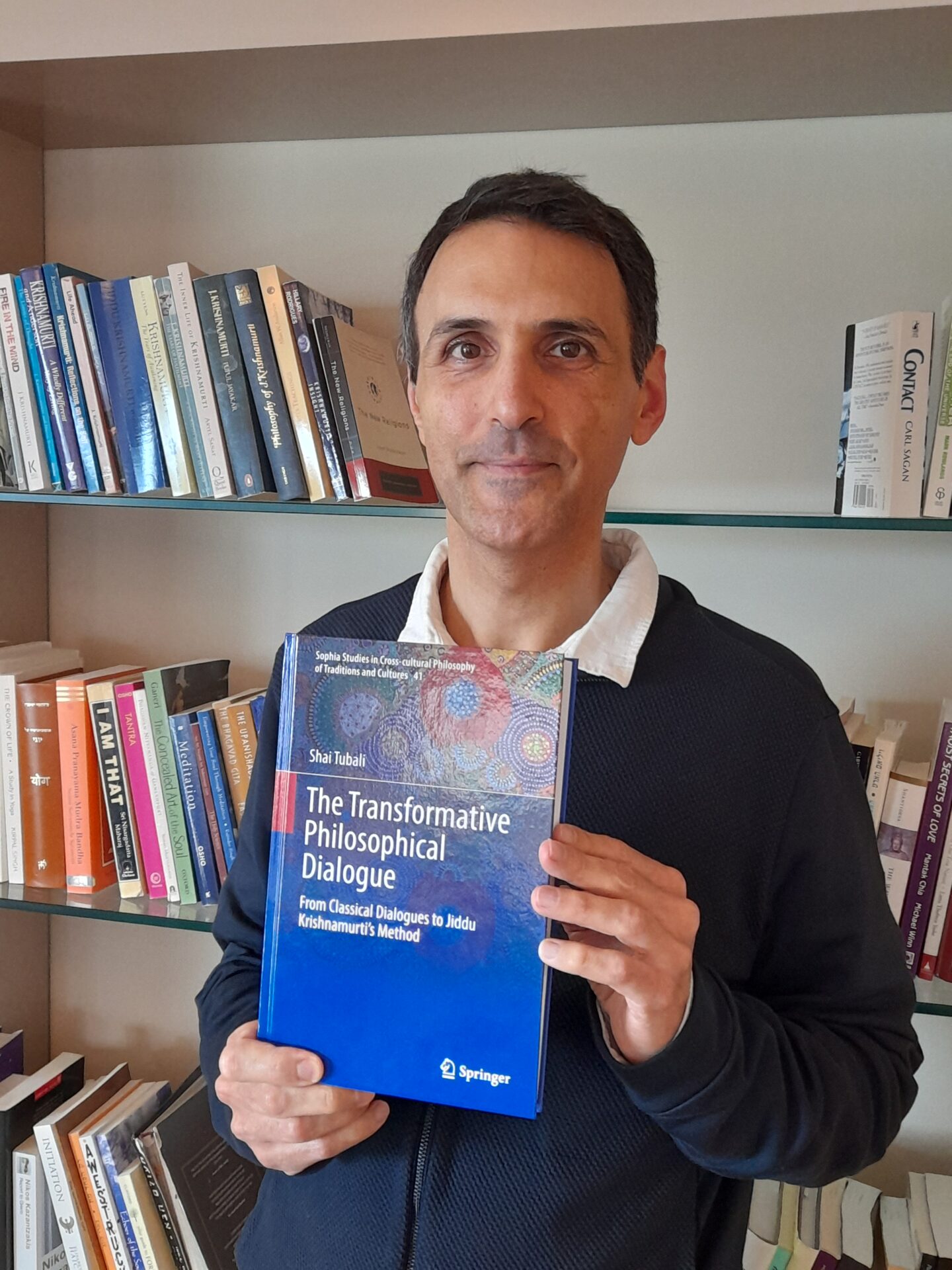 Dr Shai Tubali holding his new book. He is stood in front of a bookshelf