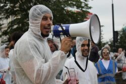 A man in a white forensic suit holding a loud speaker. He is in a crowd of people including at least one other man in a white forensic suit. Other people are indistinguishable.
