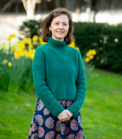 A woman wearing a green jumper and blue patterned skirt, stood on a lawn with daffodils in the background.