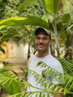 Ben is standing in amongst large green indoor plants. Ben is wearing glasses, a white t-shirt and light green baseball cap. He has a bag over his shoulder. 