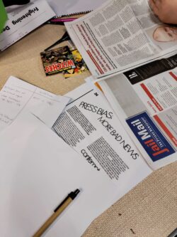 Newspapers and articles are strewn across a table. Someone's hand is in the top right hand corner of the image, poised to turn the page.