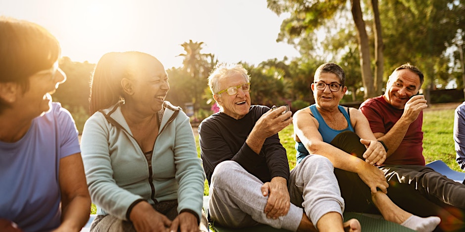 5 older people sat on the grass laughing. The reflection of the sun partially obscures the faces of the people on the left of the image