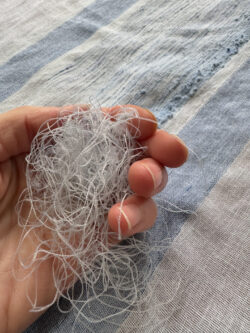 A hand cradles a tangled bundle of fine threads against a background of frayed, striped blue-and-white woven fabric, suggesting textile repair or crafting.
