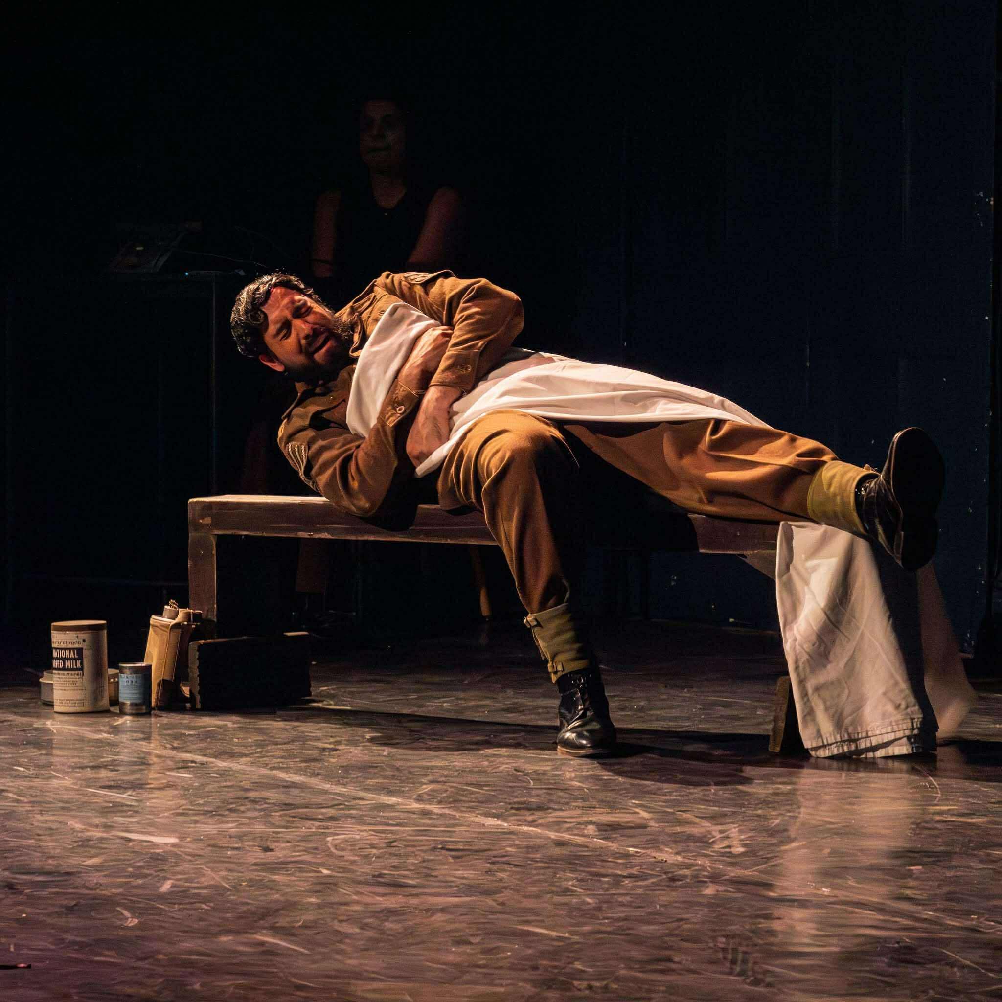A male actor in a prisoner of war costume lays on a wooden bench in pain
