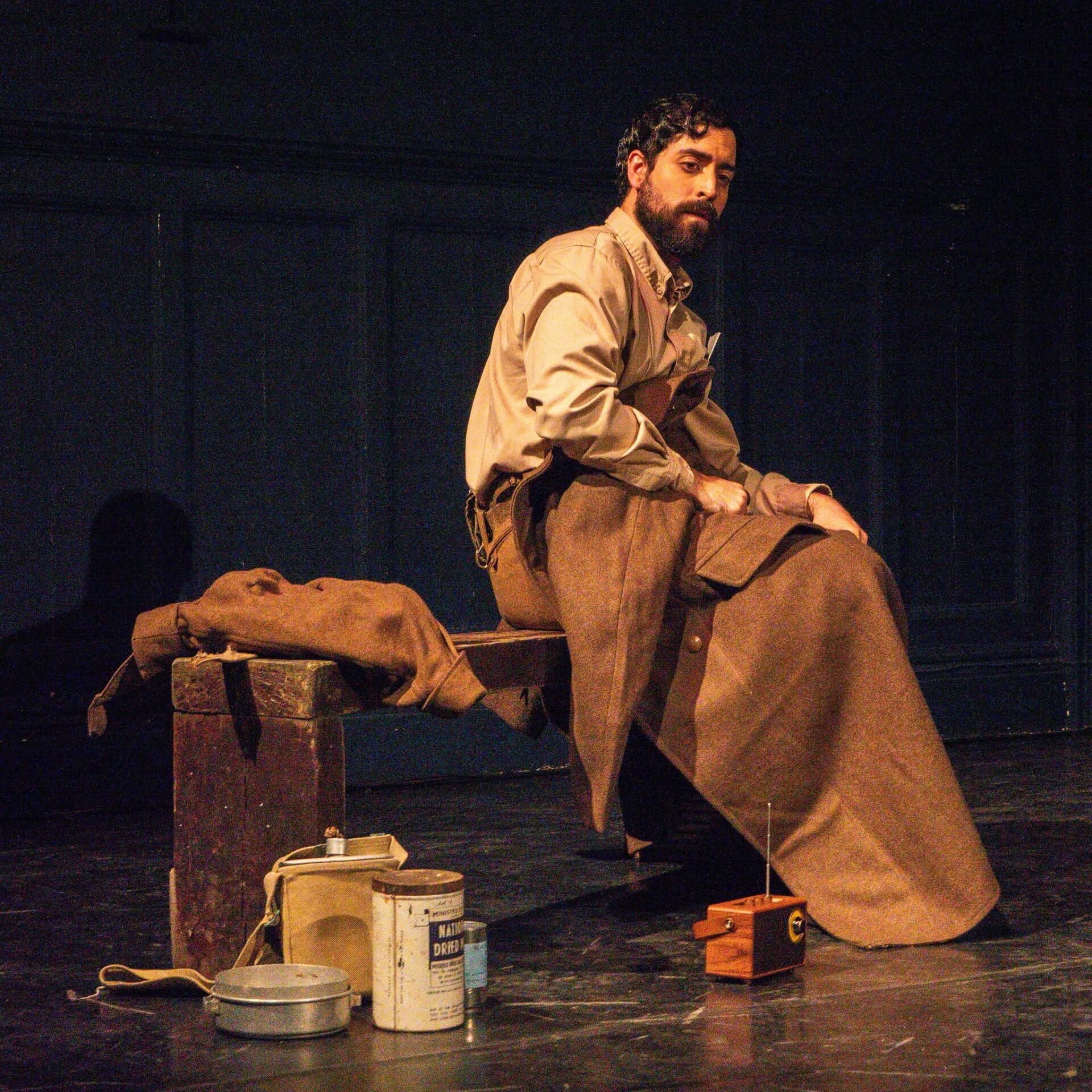 A male actor in a prisoner of war costume sits on a wooden bench, performing for the audience.